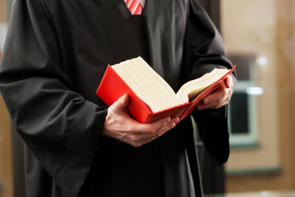 Juge ou Avocat en Robe Noire Tenant un Livre de Loi Rouge Juge en robe noire tenant un épais livre de loi rouge ouvert, symbolisant la justice et le droit.