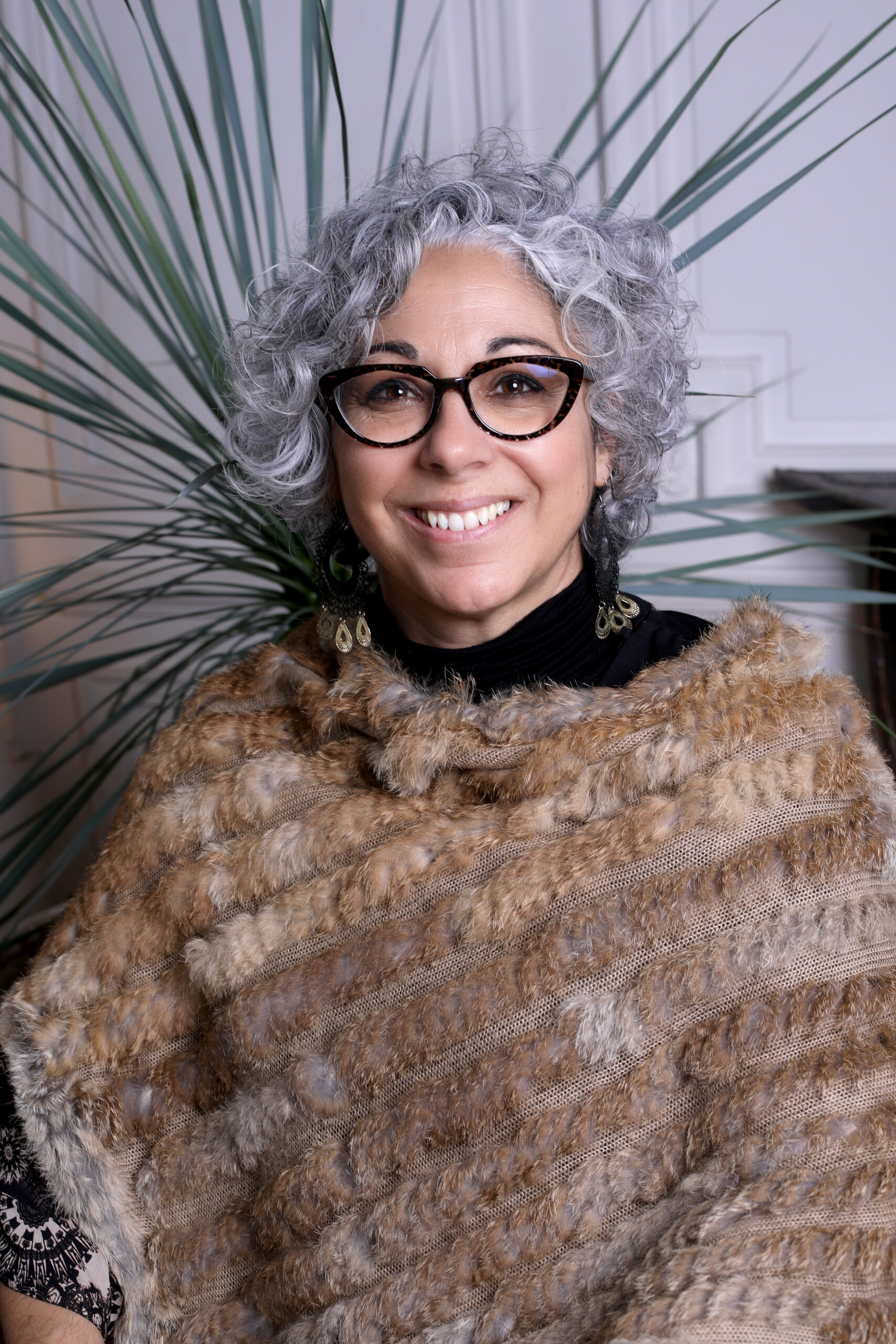 Portrait d'une femme souriante aux cheveux gris bouclés Femme souriante aux cheveux gris bouclés, lunettes écaillées et châle en fourrure devant une plante verte.