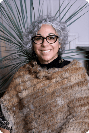 Femme souriante aux cheveux gris bouclés, lunettes noires et châle en fourrure devant une plante d'intérieur.